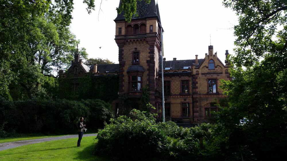 A historic building with a prominent tower, surrounded by lush greenery and a pathway.
