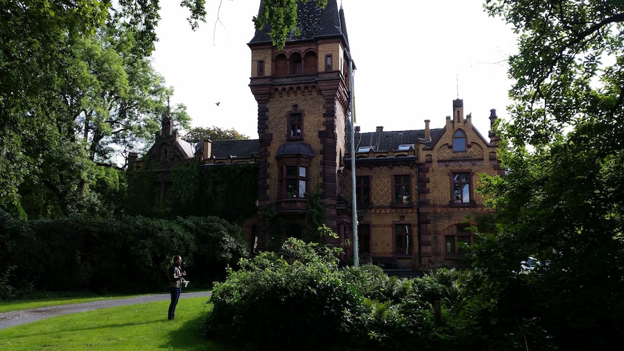 Un edificio histórico con una torre destacada, rodeado de abundante vegetación y un camino.