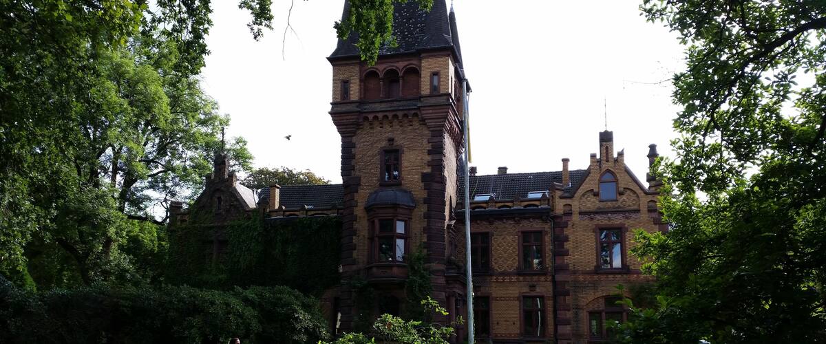 A historic building with a prominent tower, surrounded by lush greenery and a pathway.