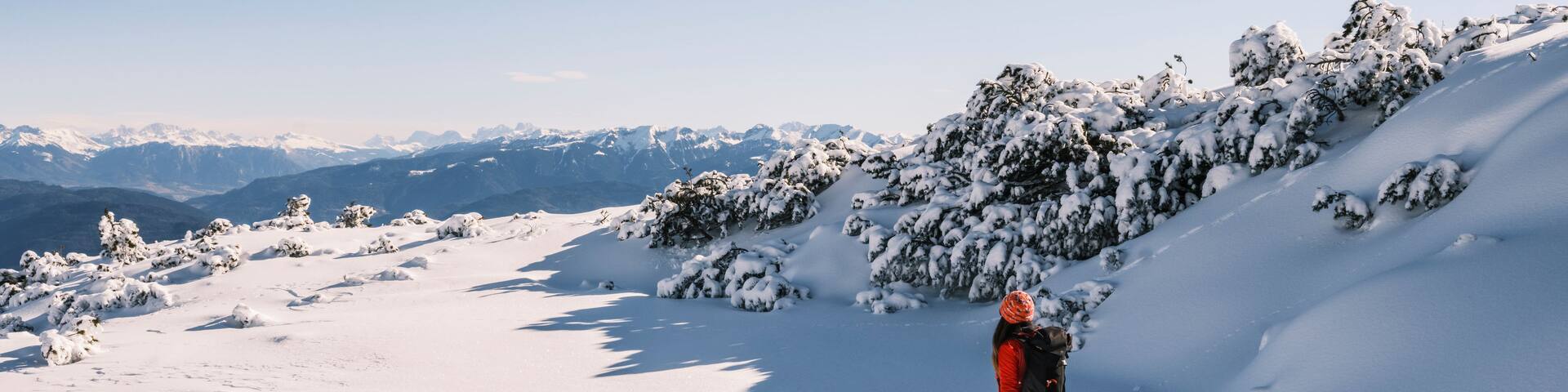 A woman snowshoes across a snowy landscape with distant mountains under a blue sky. Paganella, Roda refuge,Trentino, Italy