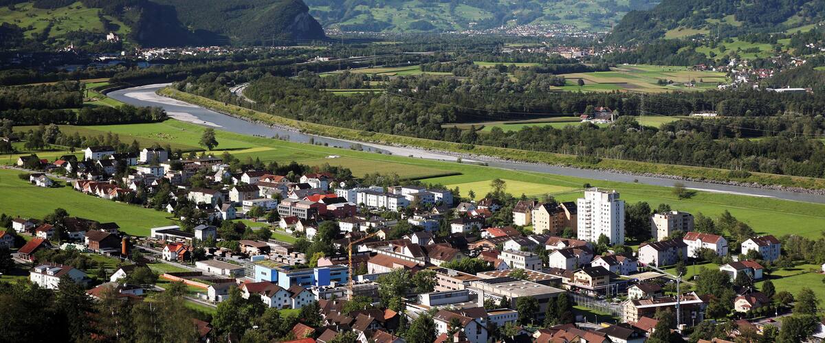 A town nestled in a valley with a river, surrounded by lush green hills and snow-capped mountains in the background.