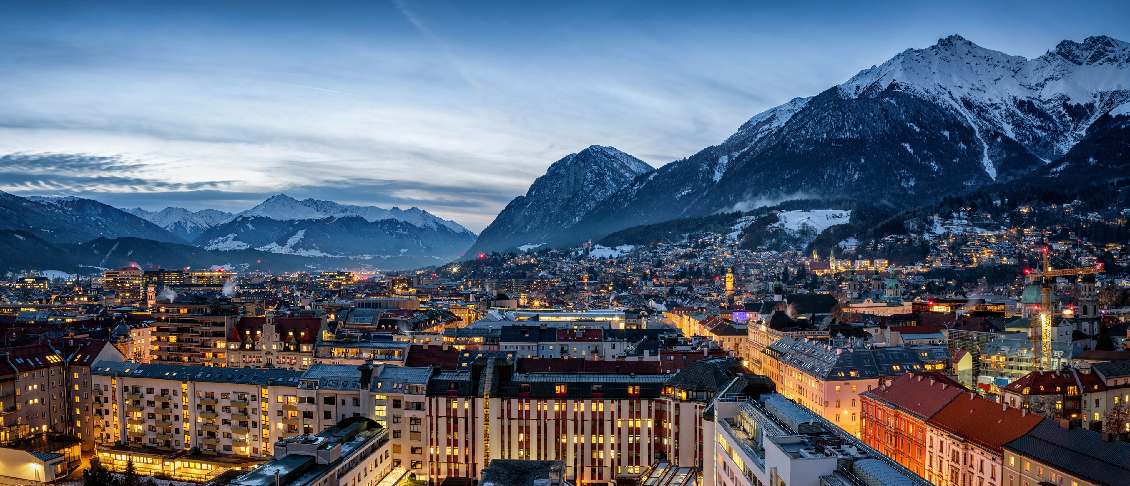 A city with snow-covered mountains in the background and illuminated buildings.