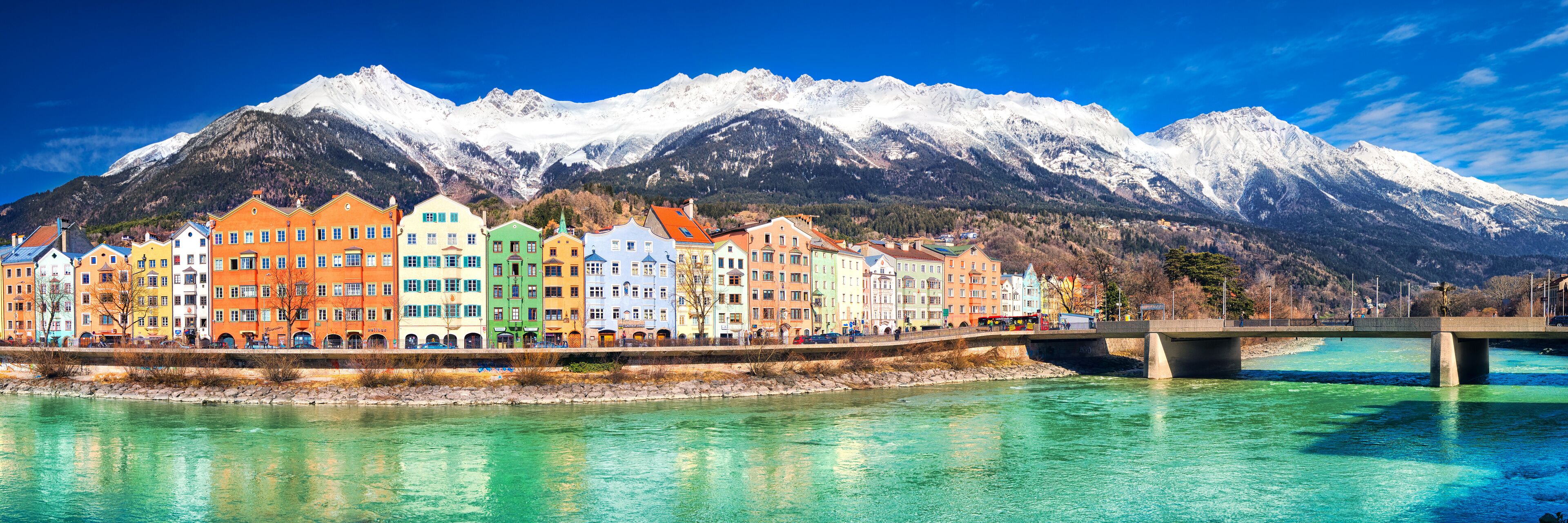 Colorful buildings along a river with a bridge and snow-capped mountains in the background.