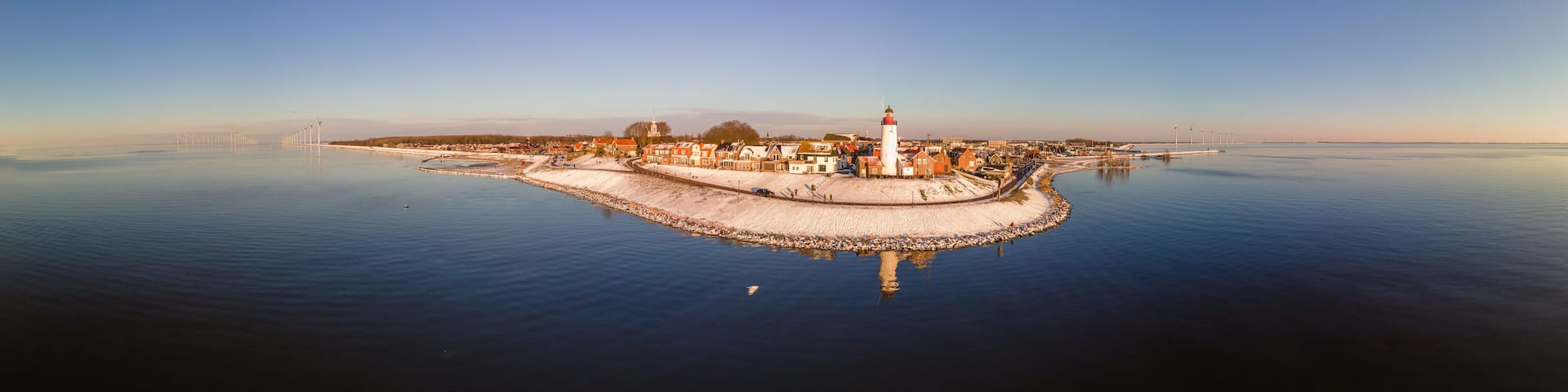 A coastal town with a prominent lighthouse, calm waters, and a snow-covered island.