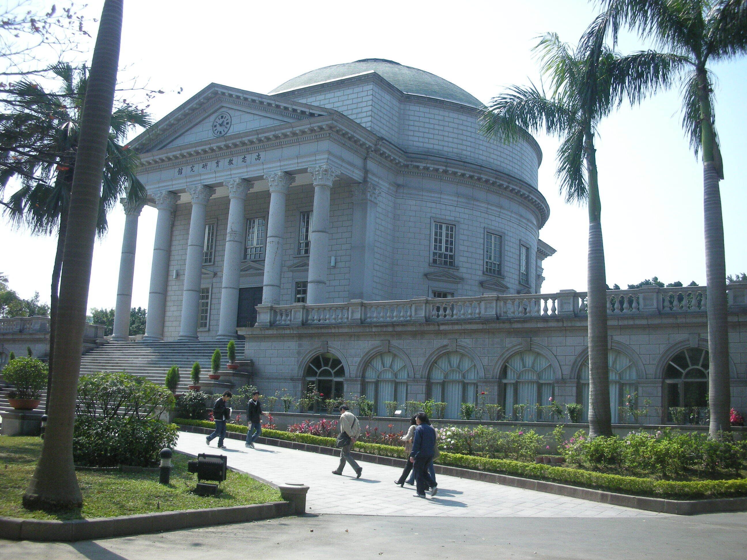 Un edificio grande de estilo clásico con columnas y una cúpula, rodeado de palmeras y un jardín cuidado.
