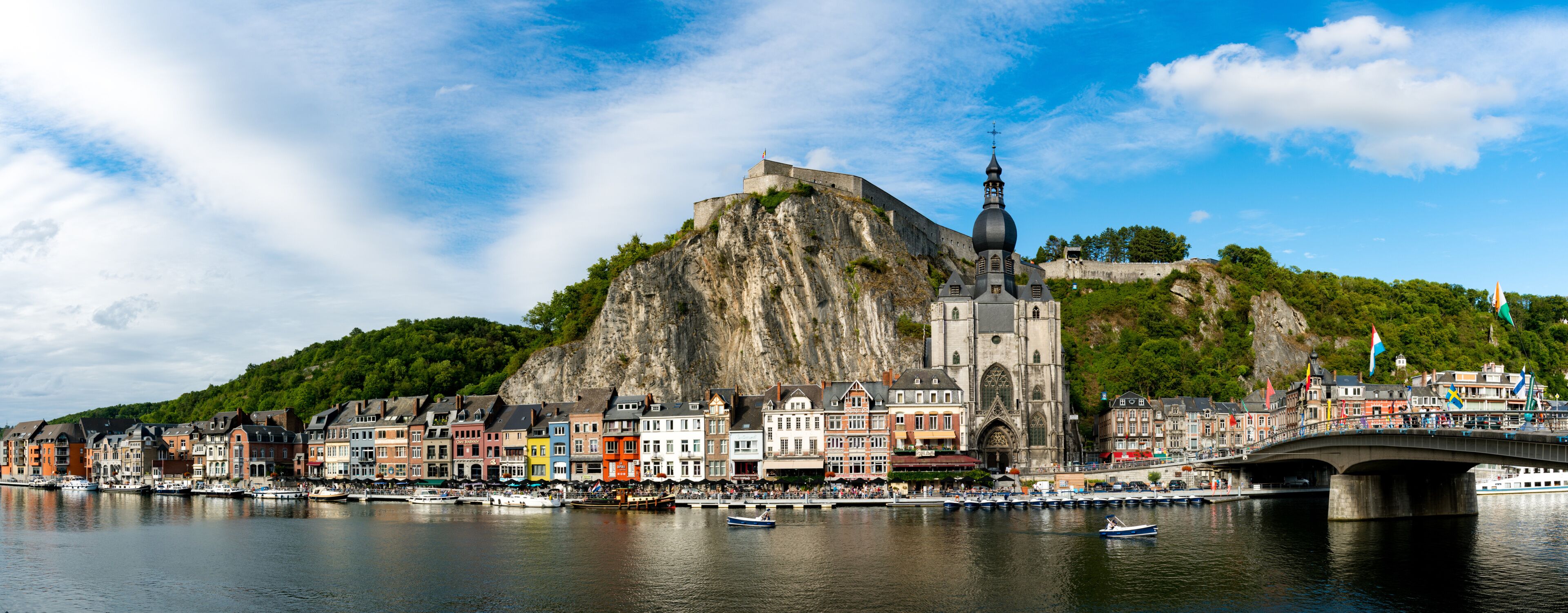 Uma cidade histórica com edifícios coloridos, uma igreja imponente, uma colina rochosa e uma ponte sobre um rio calmo.