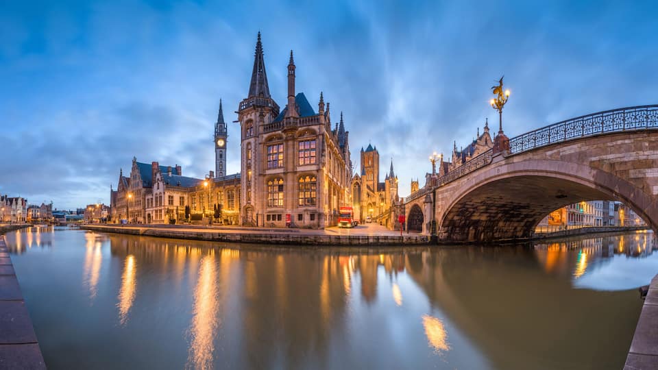 A historic bridge over a calm river, surrounded by ornate buildings and a clear evening sky.
