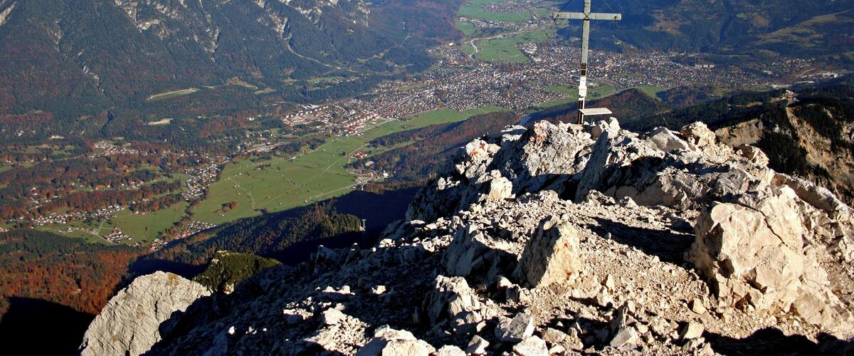 A cross on a mountain peak overlooking a valley with a town and a lake.