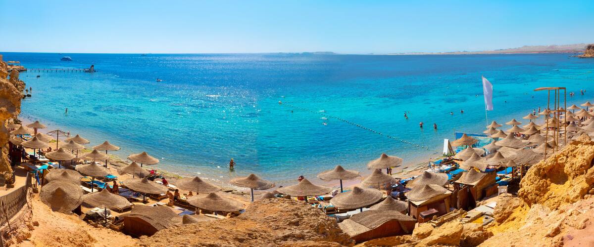 A beach with clear blue water, thatched umbrellas, and rocky cliffs.