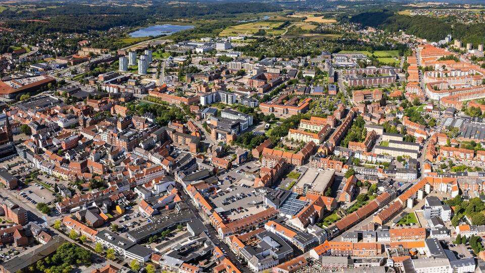 An aerial view of a densely built urban area with a mix of residential and commercial buildings, surrounded by green fields and a lake.