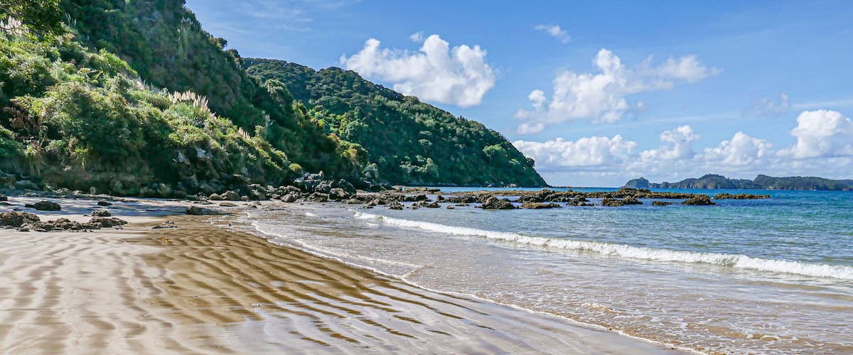 A sandy beach with gentle waves, a rocky shoreline, and a lush green hillside in the background.