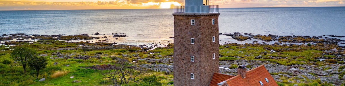 A lighthouse with a red roof, a brick tower, and a small adjacent building with a red roof, set against a backdrop of a rocky shoreline and a sunset over the ocean.