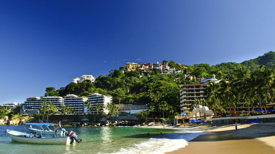 A beach with a boat, palm trees, and buildings on a hillside.