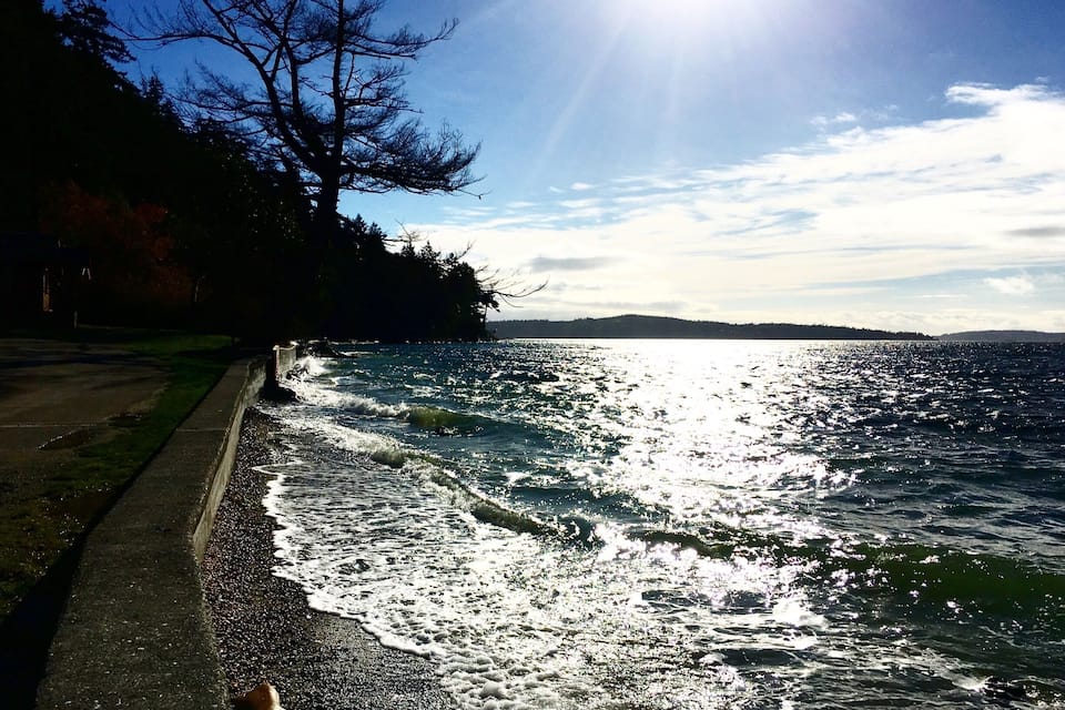 A coastal road with waves crashing onto the shore, a clear sky, and a distant shoreline with trees.