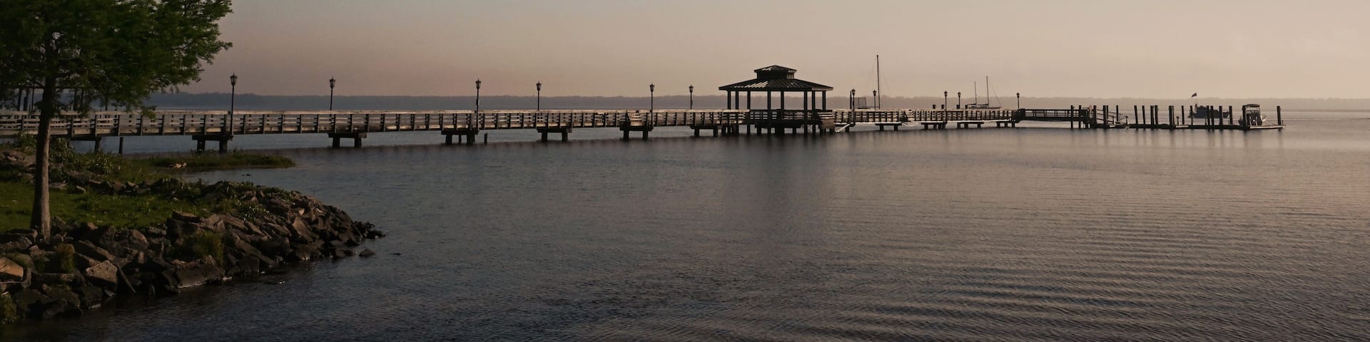 A calm lake with a wooden pier extending into the water, a gazebo at the end, and a tree on the left.