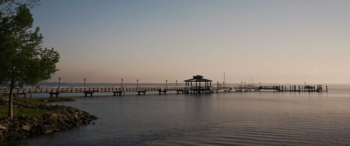 A calm lake with a wooden pier extending into the water, a gazebo at the end, and a tree on the left.