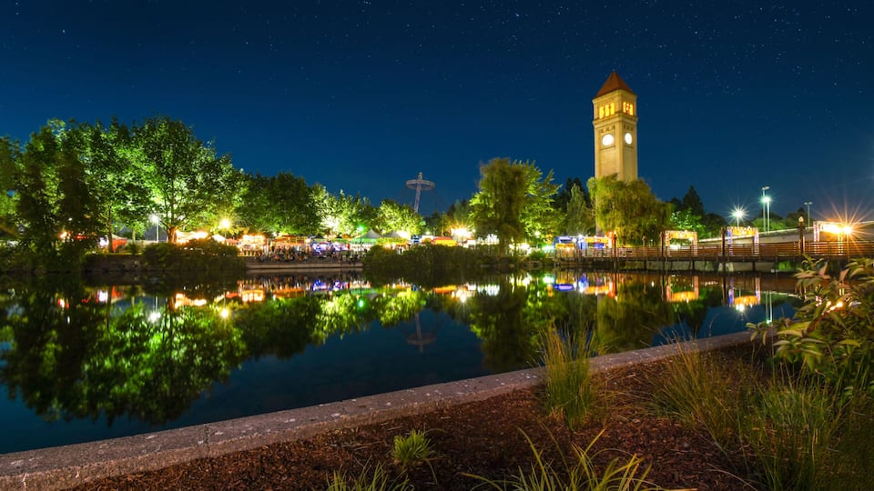 A nighttime view of a park with a tower, trees, and a calm lake reflecting lights.