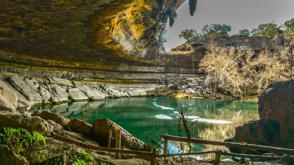 A natural cave pool with clear water, surrounded by rocky terrain and sparse vegetation.