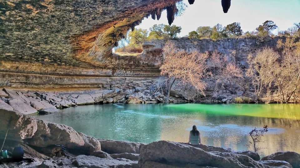 A cave entrance with stalactites, a clear green pool, and rocky terrain.