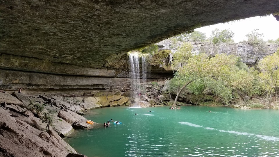 A waterfall under a rock overhang with clear water and people swimming.