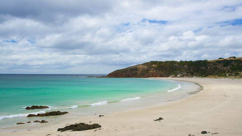A beach with clear turquoise waters, a sandy shore, and a rocky outcrop with vegetation on a hillside.