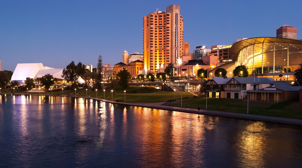 Un paisaje urbano al atardecer con un edificio moderno destacado, un río que refleja las luces y un parque con árboles.