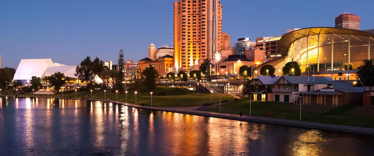 Un paisaje urbano al atardecer con un edificio moderno destacado, un río que refleja las luces y un parque con árboles.