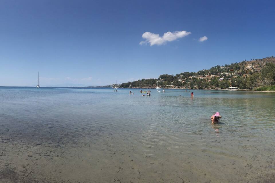 Ein ruhiger Strand mit klarem Wasser, ein entfernter Segelboot und eine hügelige Landschaft mit Bäumen.