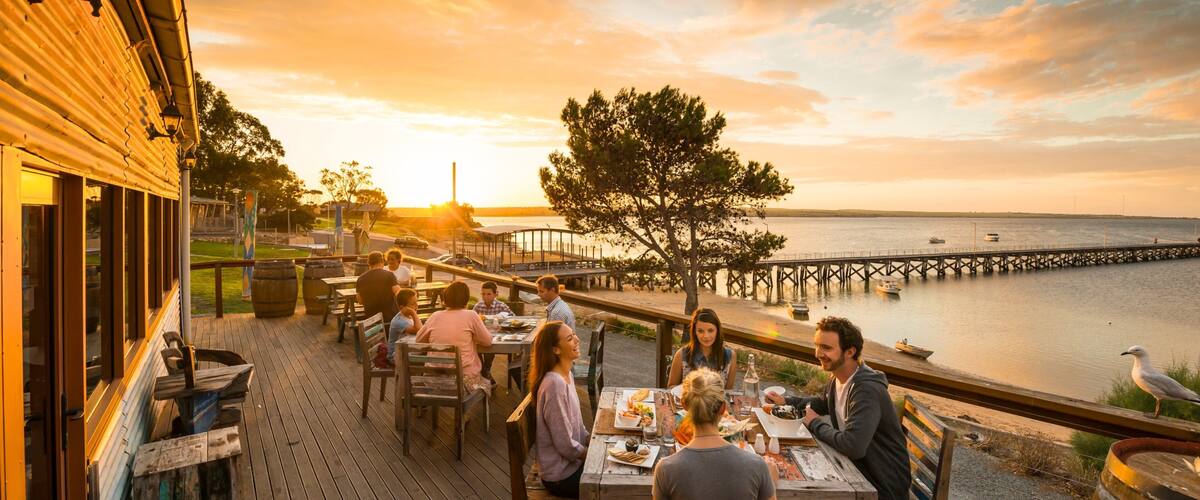 A wooden deck with people dining at tables overlooking a body of water at sunset.