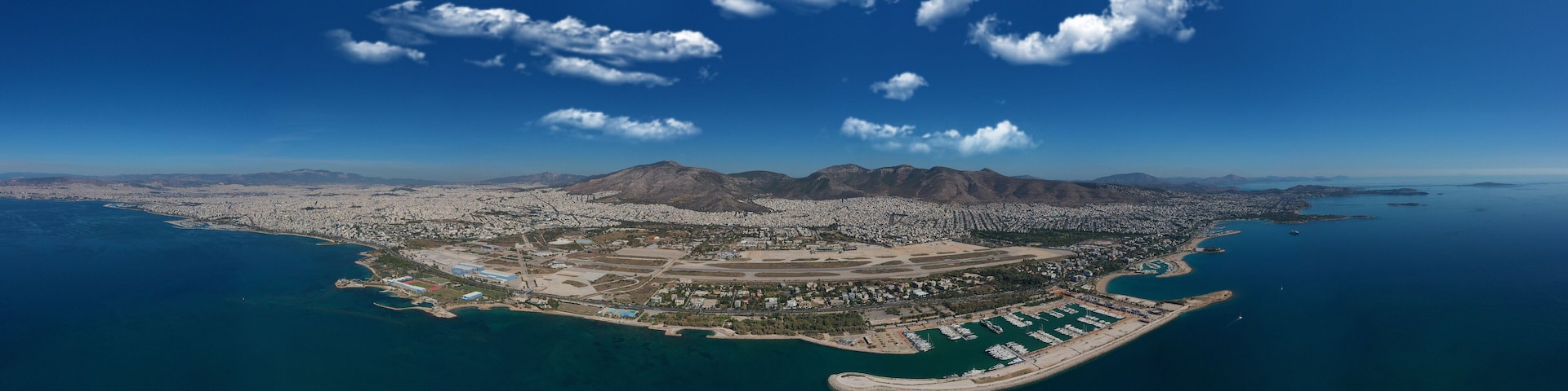 An aerial view of a coastal city with a mountainous backdrop, a marina, and a large airport.