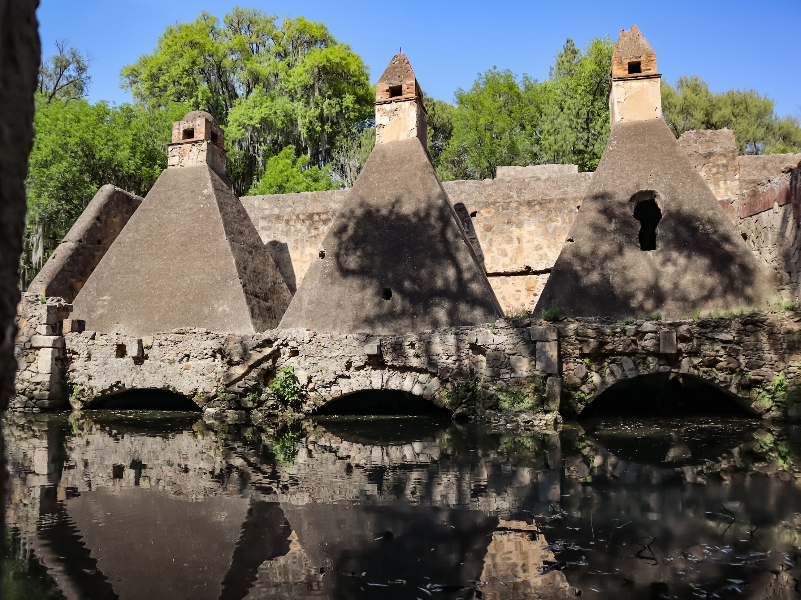 Antiguas estructuras de piedra con techos cónicos y un puente de piedra sobre una superficie acuática que refleja el entorno.