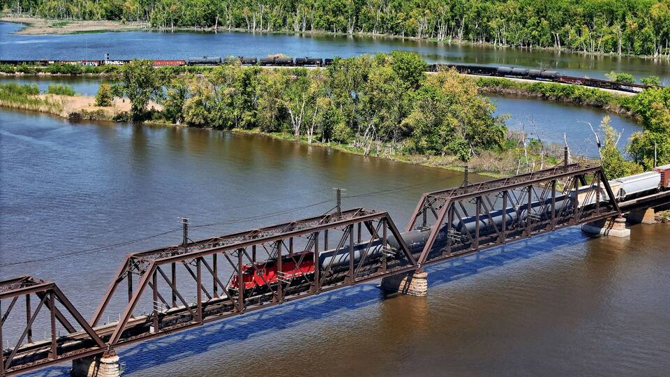 A train crossing a river over a steel bridge with a forested landscape in the background.