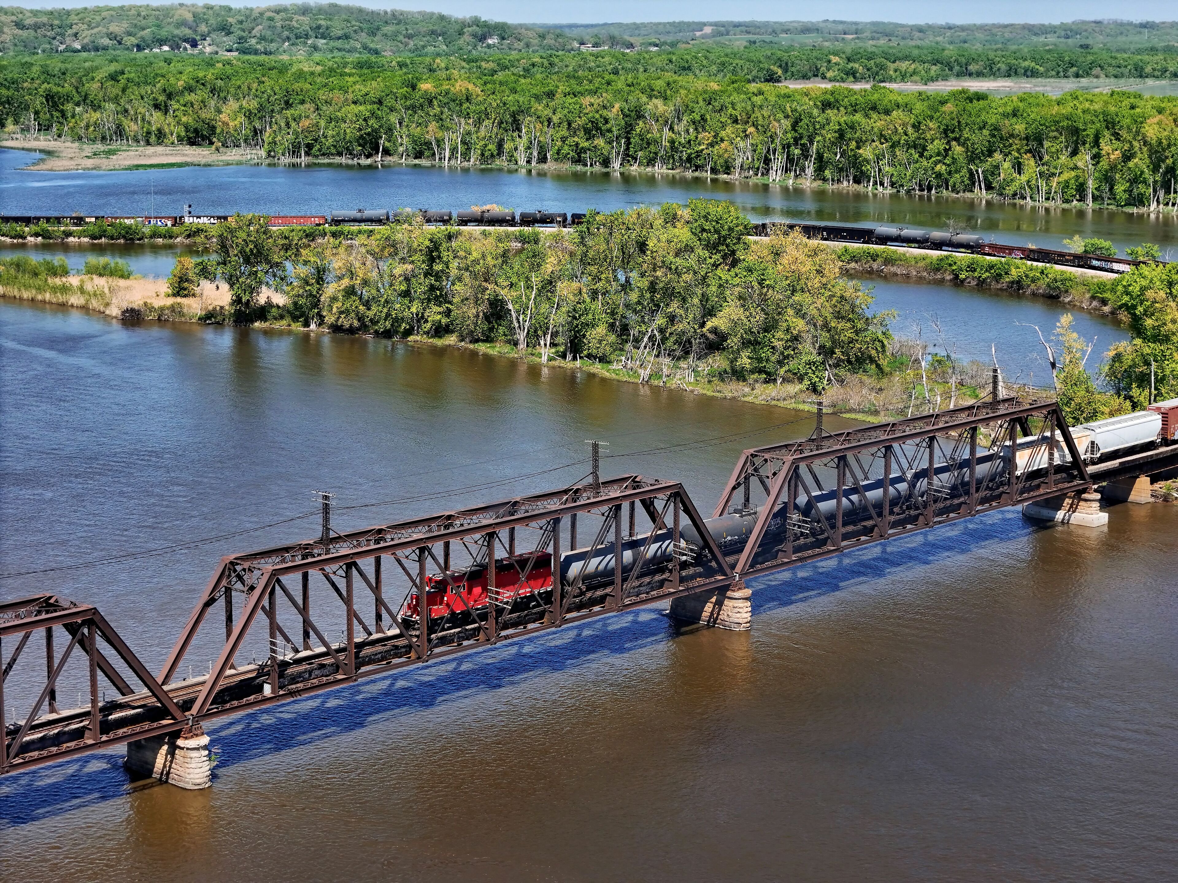 A train crossing a river over a steel bridge with a forested landscape in the background.