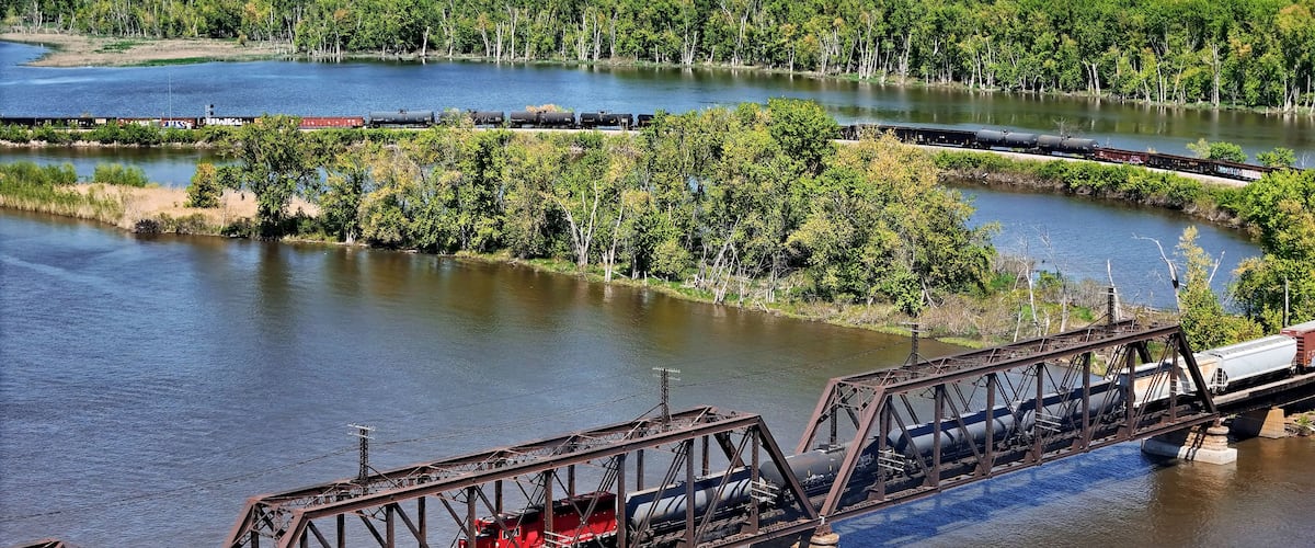 A train crossing a river over a steel bridge with a forested landscape in the background.