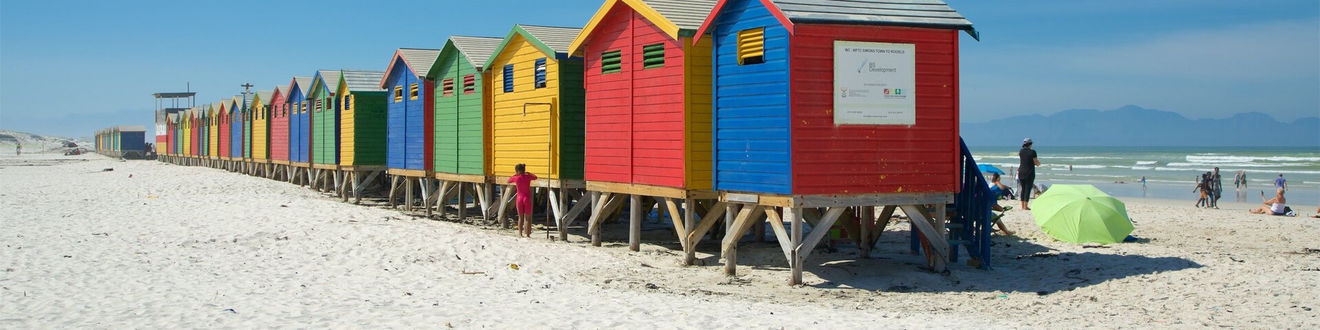 Una hilera de cabañas de playa coloridas sobre pilotes a lo largo de una playa arenosa bajo un cielo despejado.
