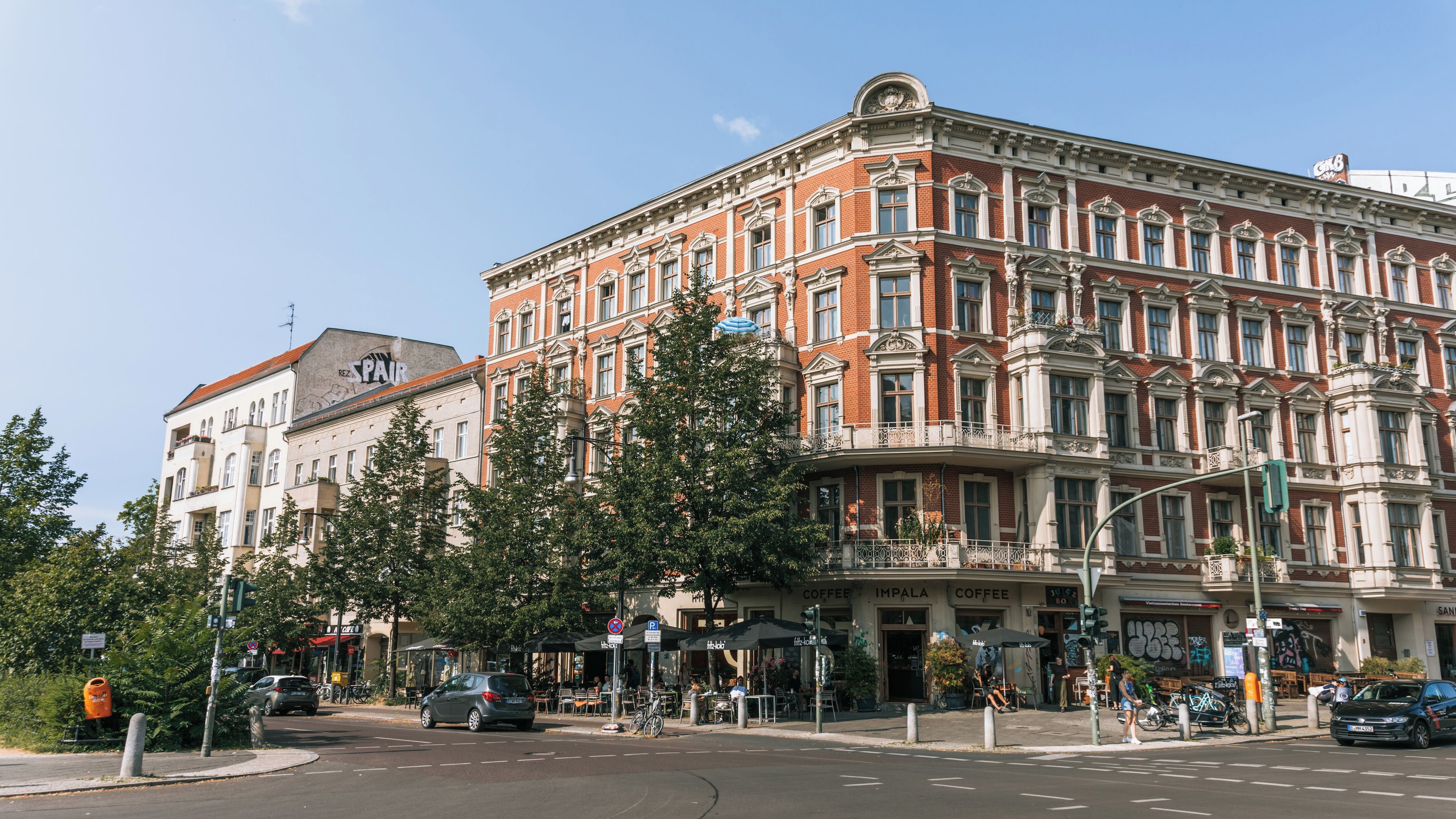 Un bâtiment historique à l’architecture riche et élaborée, une rue animée par la circulation des voitures et des piétons, et une terrasse de café.