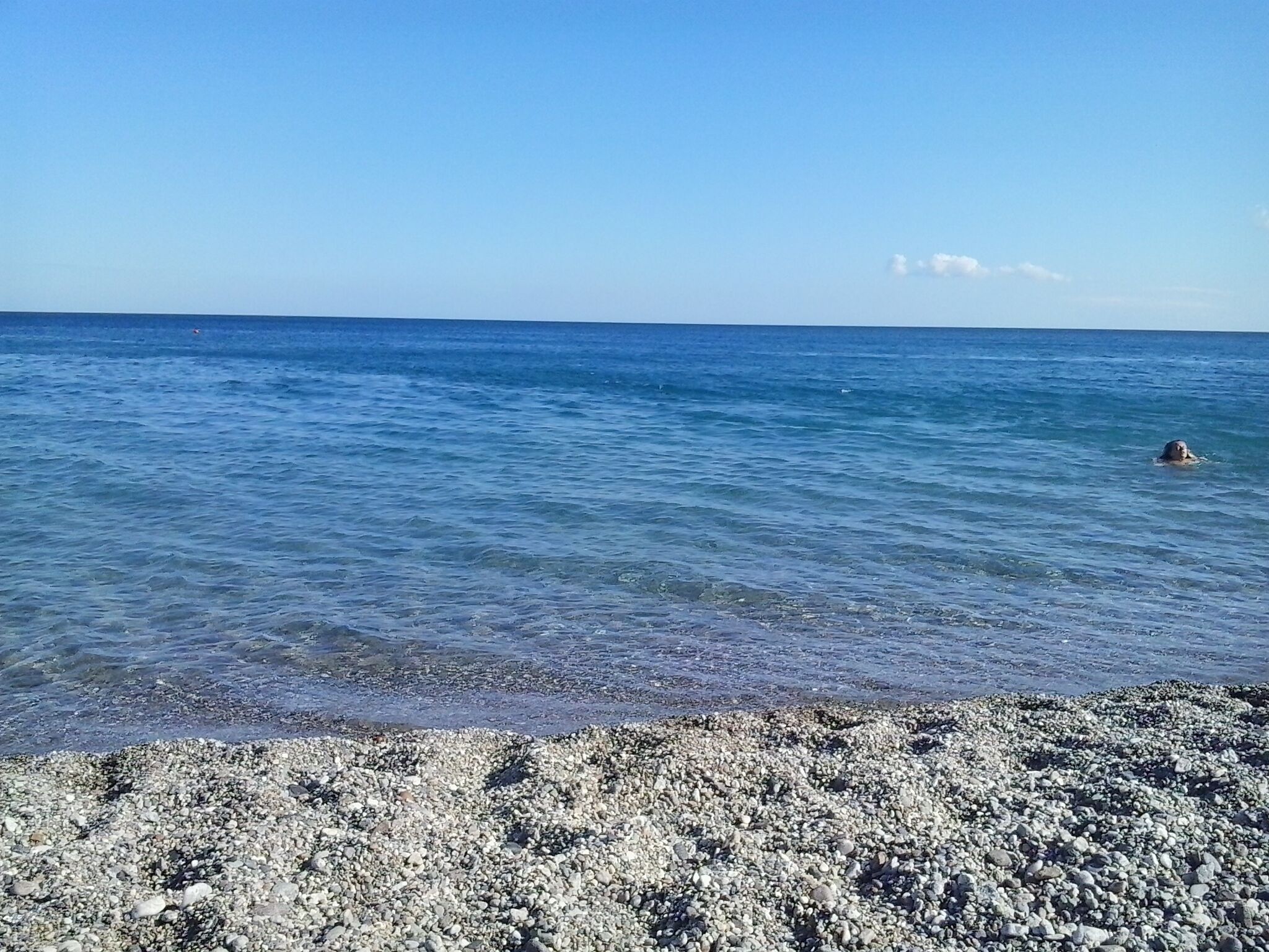A pebble beach with calm sea and a swimmer in the distance.