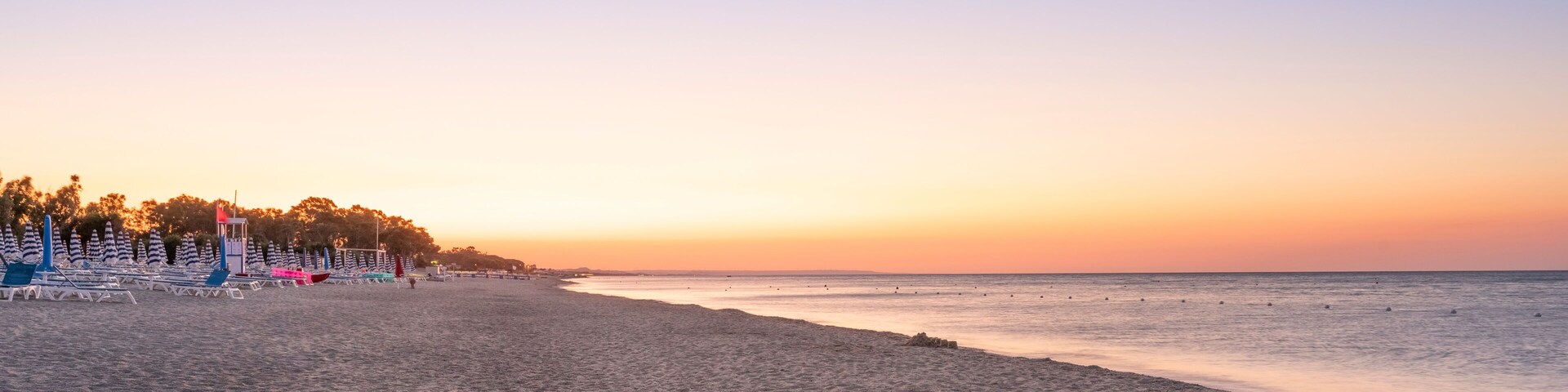 A beach with lounge chairs and umbrellas, a lifeguard tower, and a sunset over the ocean.