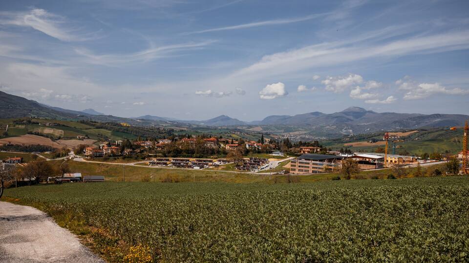 A town nestled in a valley with mountains in the background, surrounded by fields and construction cranes.