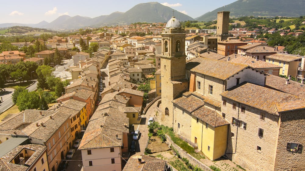 A historic town with a prominent bell tower, cobblestone streets, and surrounding mountains.