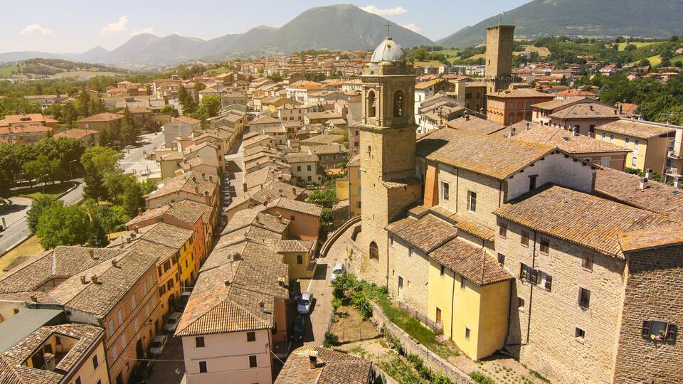 A historic town with a prominent bell tower, cobblestone streets, and surrounding mountains.