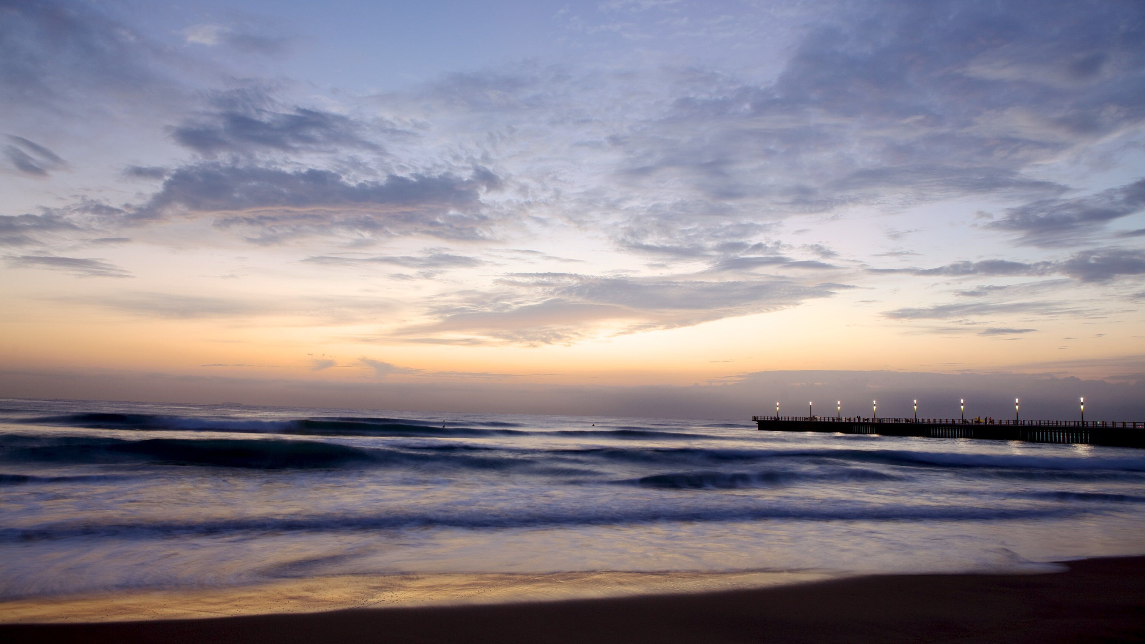 A beach with waves and a pier with lights.
