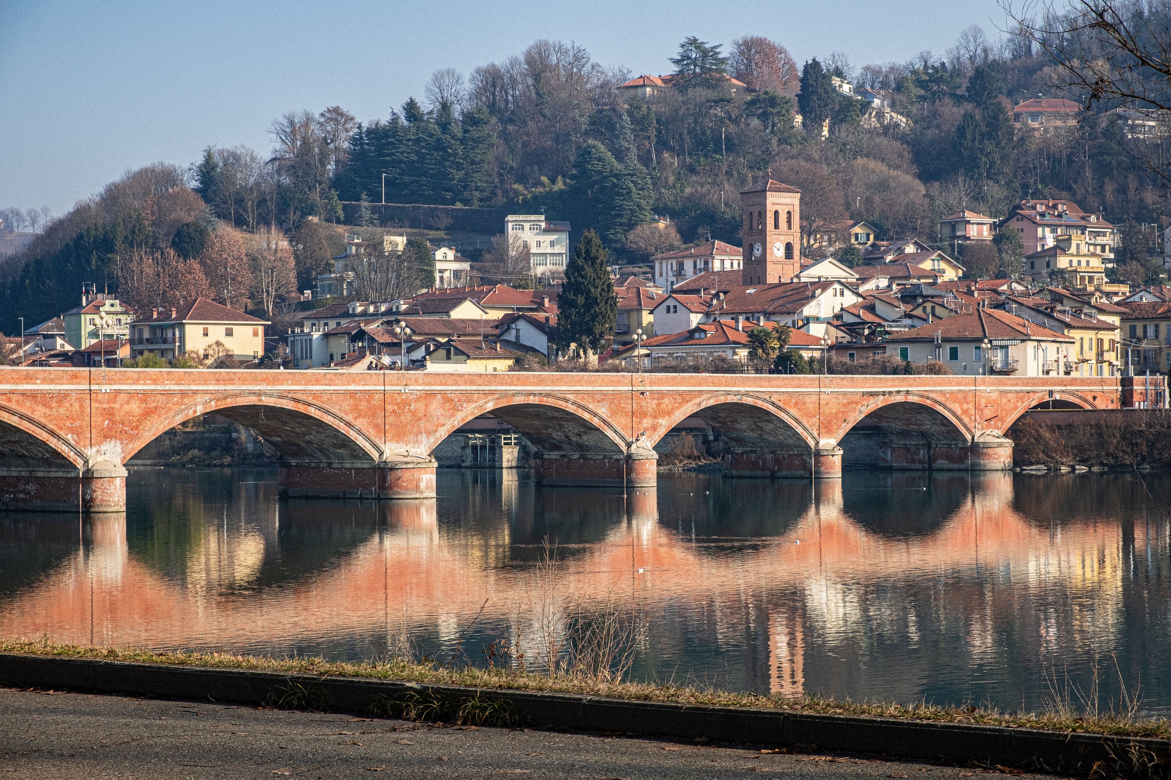 A stone bridge over a calm river, with a town and forested hills in the background.
