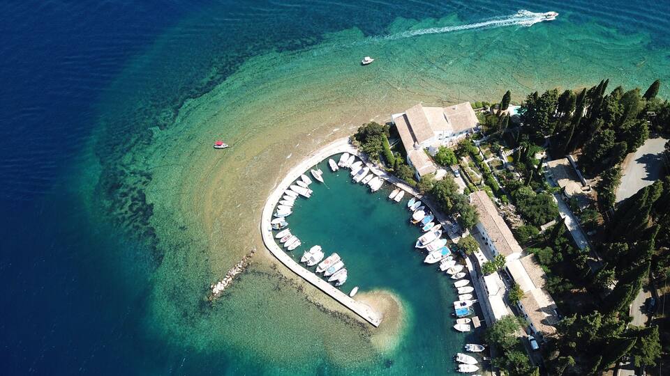 An aerial view of a coastal area with clear turquoise waters, a marina with boats, and a small building surrounded by trees.