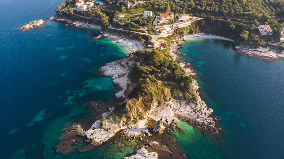 An aerial view of a coastal area with clear turquoise waters, rocky outcrops, and scattered buildings.