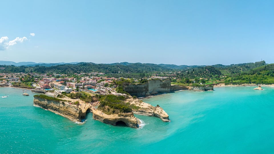 A coastal town with a prominent rocky peninsula, clear turquoise waters, and a backdrop of green hills.