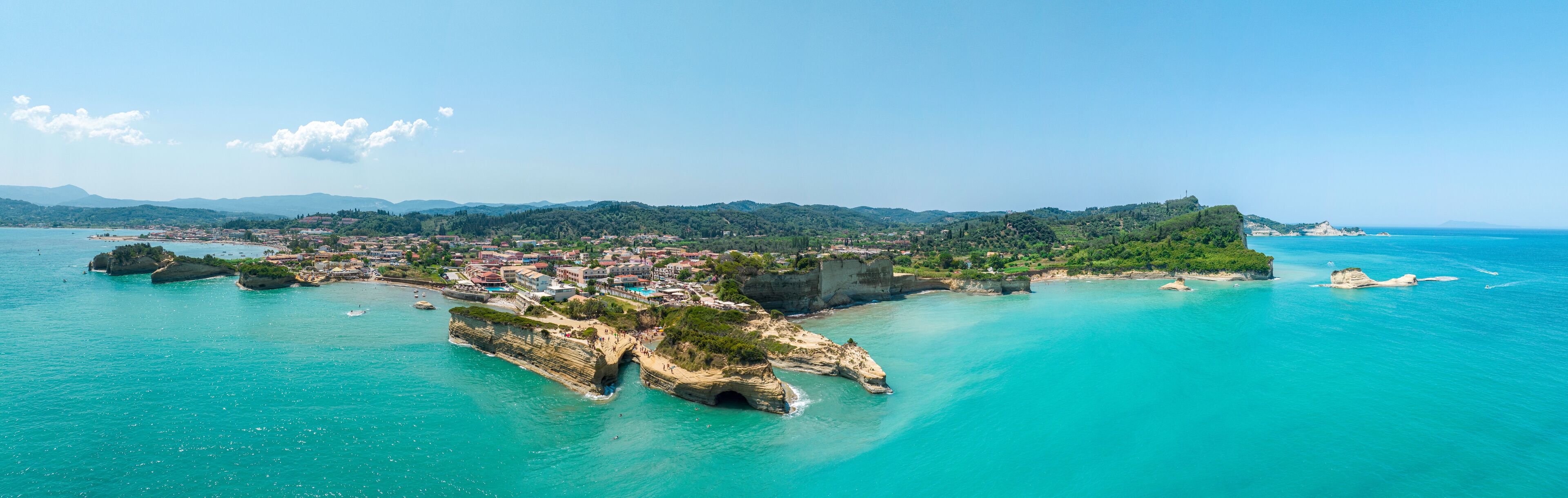 A coastal town with a prominent rocky peninsula, clear turquoise waters, and a backdrop of green hills.