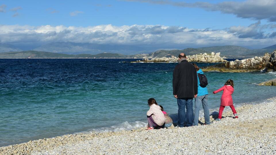 A rocky shoreline with clear blue water, a distant mountain range, and a few people by the water.
