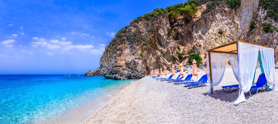 A beach with clear turquoise water, a rocky cliff, and blue lounge chairs.