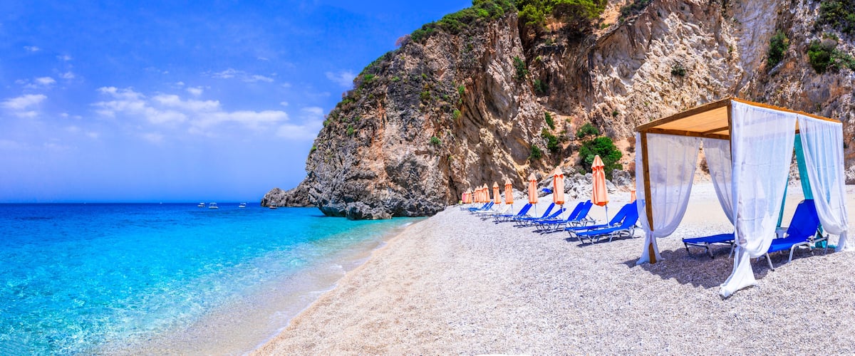 A beach with clear turquoise water, a rocky cliff, and blue lounge chairs.